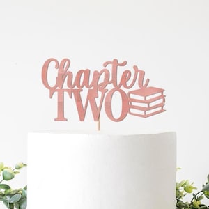 May include: A rose gold cake topper reading "Chapter TWO" with a book stack design. The topper is placed on a white cake, with green foliage in the foreground. Ideal for a second birthday or anniversary celebration.