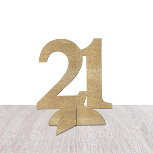 May include: A gold-colored wooden number 21, standing upright on a light-colored wooden surface. The number is cut out and has a textured appearance. The background is plain white.