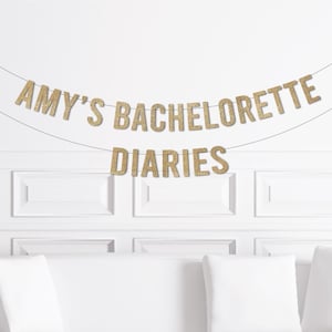 May include: A gold glitter banner that reads "AMY'S BACHELORETTE DIARIES" hangs against a white wall. The banner is strung on a thin string. Below the banner is a white couch with white pillows.