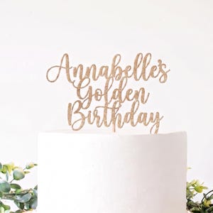 May include: A gold glitter cake topper featuring the text Annabelle's Golden Birthday in a cursive script font. The topper is mounted on a thin wooden stick and is positioned on top of a plain white cake.