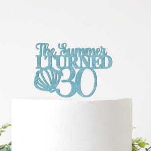 May include: A blue glitter cake topper with the text "The Summer I Turned 30" and a seashell design. The topper is on a white cake, with green foliage in the background.