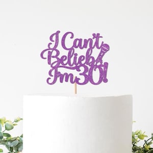 Peut inclure: Un cake topper violet pailleté avec l'inscription "I Can't Believe I'm 30 !" et un motif de microphone. Le topper est posé sur un gâteau blanc, avec des plantes vertes en arrière-plan. Il est soutenu par un bâton en bois.