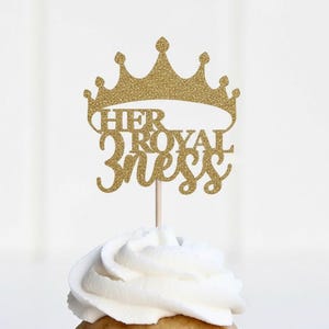 May include: A gold glitter crown cake topper with the words "HER ROYAL 3NESS" in a cursive font. The topper is attached to a wooden stick and placed on top of a cupcake with white frosting. The background is a soft, blurred white.