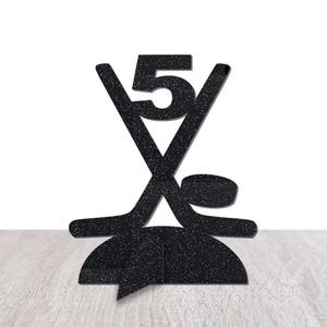 May include: Black hockey-themed cake topper with the number 5. The design features crossed hockey sticks, a puck, and a base. The topper is made of a textured material and is suitable for a birthday or sports-themed celebration.