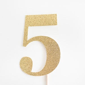 以下が含まれることがあります： 金色のグリッターでできた数字5のケーキトッパー。数字は金色のグリッターの厚紙から切り抜かれ、木製の棒につけられています。