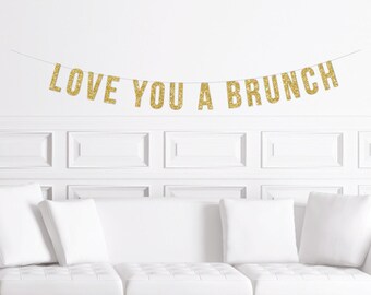 May include: A gold glitter banner that reads "LOVE YOU A BRUNCH" is strung across a white wall. Below the banner is a white couch with white pillows. The banner is a festive decoration for a brunch event.