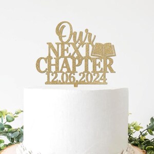 以下が含まれることがあります： 「Our Next Chapter」と日付「12.06.2024」の文字が入った、金色のグリッターケーキトッパー。トッパーには開いた本のデザインが含まれています。ケーキは白く、緑の植物で飾られています。
