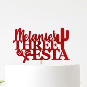 May include: A red glitter cake topper with the words "Melanie's THREE FIESTA" and maracas and a cactus design. The topper is on a white cake, perfect for a birthday celebration.