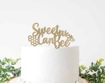 May include: A gold glitter cake topper with the phrase "Sweet as Can Bee" in cursive script. The design includes a honeycomb and a bee. The topper is placed on top of a white cake, with greenery in the background.