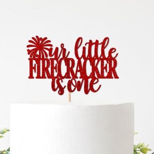May include: A red glitter cake topper with the words "Your little firecracker is one" on a white cake. The topper is on a wooden stick, set against a white background with greenery.