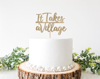 May include: A white cake with a gold glitter cake topper that reads "It Takes a Village." The cake sits on a wooden cake stand and is decorated with white frosting. Greenery surrounds the cake, creating a celebratory atmosphere.