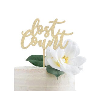 May include: A gold glitter cake topper with the words "lost count" in a cursive font. The topper is on a cake decorated with a white flower and green leaves. The cake has a white frosting.