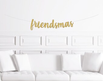 May include: Gold glitter "Friendsmas" banner hanging on a white wall above a white couch with white pillows.
