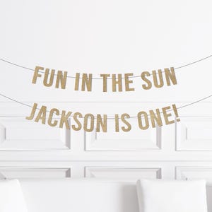 May include: Gold glitter banner with the text "FUN IN THE SUN" and "JACKSON IS ONE!" strung across a white wall. The banner is a festive decoration, perfect for a birthday celebration.