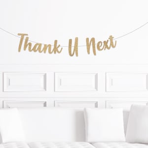 Peut inclure: Une bannière dorée à paillettes avec l'inscription "Thank U Next" est suspendue sur un mur blanc. En dessous, un canapé blanc avec des coussins blancs est visible. Le mur présente un motif de panneaux décoratifs.