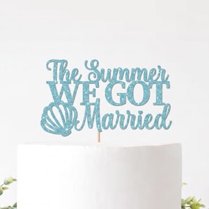 May include: A light blue glitter cake topper featuring the text The Summer WE GOT Married with a seashell graphic on the left side. The topper is mounted on a thin stick and is positioned on top of a plain white cake surface.