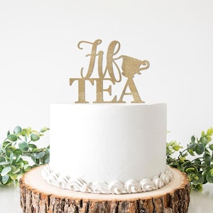 May include: A white cake with a decorative gold glitter cake topper that reads "Tea". The cake sits on a wooden base and is surrounded by greenery. The cake topper features a teacup design.