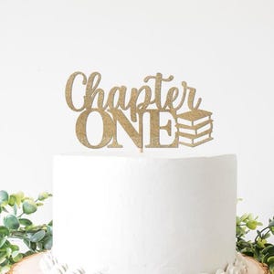 May include: A gold glitter cake topper that reads "Chapter ONE" with a stack of books. The topper is placed on top of a white cake, with greenery in the foreground. Perfect for a first birthday or milestone celebration.