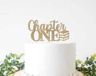 Pode incluir: Um topo de bolo dourado com glitter que diz "Chapter ONE" com uma pilha de livros. O topo está colocado em cima de um bolo branco, com vegetação em primeiro plano. Perfeito para um primeiro aniversário ou celebração.