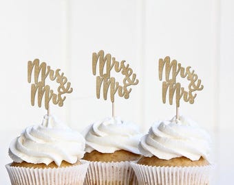 May include: Three cupcakes with white frosting and gold glitter "Mrs & Mrs" toppers. The cupcakes are in white paper liners, and the background is a plain white surface.