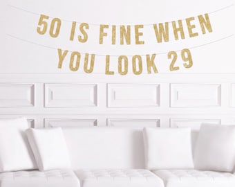 May include: Gold glitter banner with the text "50 IS FINE WHEN YOU LOOK 29" hanging against a white wall. Below the banner is a white couch with white pillows.