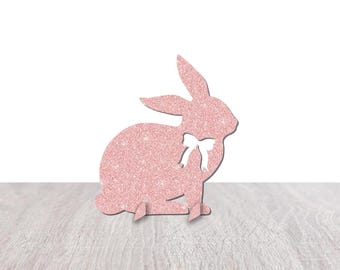 May include: A pink glitter bunny rabbit cutout with a white bow. The bunny is sitting on a light wood surface.