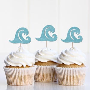 Peut inclure: Trois cupcakes avec glaçage blanc et des décorations en forme de vagues bleu clair. Les cupcakes sont dans des caissettes en papier blanches et reposent sur une surface blanche. Les décorations en forme de vagues ajoutent une touche décorative.
