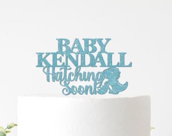 Puede incluir: Un adorno para tarta de purpurina azul claro con las palabras "BABY KENDALL Hatching Soon" y una silueta de dinosaurio. El adorno está colocado encima de una tarta blanca, ideal para un baby shower.
