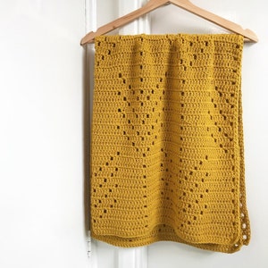 May include: A mustard yellow crocheted blanket with a diamond pattern hangs on a wooden hanger against a white wall.