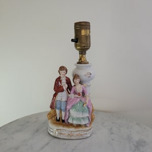 May include: A porcelain lamp base featuring a couple in 18th-century dress. The woman is wearing a pink dress and the man is wearing a red jacket and blue trousers. The lamp base is decorated with gold accents.