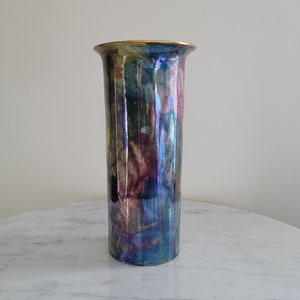 Puede incluir: Un jarrón cilíndrico alto con un borde dorado y un esmalte iridiscente de colores. El esmalte presenta tonos de azul, morado, verde y rosa.