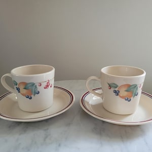 May include: Two white ceramic coffee cups with a fruit design and red, blue, and green accents. Each cup is on a white saucer with a red and blue trim.