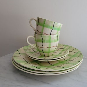 May include: A set of three green and white checked ceramic teacups and saucers. The cups and saucers have a green and white checked pattern with black and brown lines.