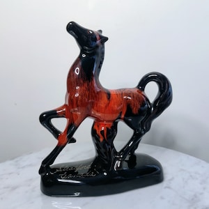 Puede incluir: Una figurita de caballo de cerámica negra y naranja con una base negra. La base tiene el texto "Stafford Canada" inscrito en ella.