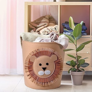 May include: A brown lion-shaped laundry basket with a white face and black eyes sits on a wooden floor. The basket is in front of a wooden shelf unit with a cactus in a pot on the top shelf. The shelf unit is against a pink wall.