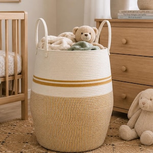 May include: A large, round woven storage basket with cream and mustard yellow accents. The basket is filled with blankets and a teddy bear. The basket has two handles and is sitting on a woven rug. A framed print that says "you are so loved" is on the wall.
