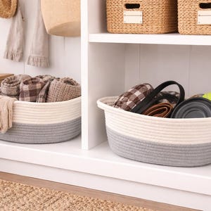 May include: Two-tone rope baskets in a white cabinet. One basket contains folded blankets and gloves. The other basket holds a black pet carrier, a wallet, a collapsible bowl, and a tennis ball. Woven baskets sit on a shelf above.