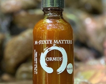 May include: A small amber glass bottle with a dark gray lid. The bottle is labeled "M-State Matters" and features a white circle with the word "ORMUS" inside. The bottle is sitting on a rock.