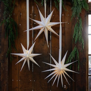 May include: Three white paper star lanterns with multiple pointed tips hanging from long cream-colored ribbons. Each star features small perforated holes that allow warm light to glow from within, creating a bright, illuminated display.