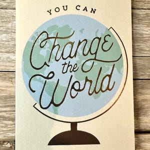 K&ouml;nnte beinhalten: Rechteckige Karte mit den Worten "YOU CAN Change the World" in goldener Schrift &uuml;ber einer hellblauen Globusillustration. Die Karte hat einen cremefarbenen Hintergrund und eine dunkelbraune Basis.