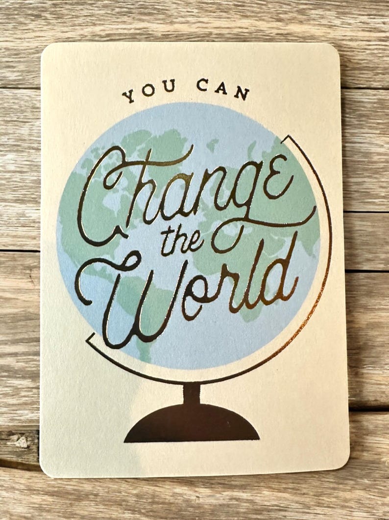 K&ouml;nnte beinhalten: Rechteckige Karte mit den Worten "YOU CAN Change the World" in goldener Schrift &uuml;ber einer hellblauen Globusillustration. Die Karte hat einen cremefarbenen Hintergrund und eine dunkelbraune Basis.