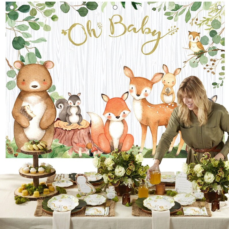 May include: A woodland-themed baby shower setup with a backdrop featuring watercolor illustrations of forest animals and the text "Oh Baby." The table is set with plates, napkins, and floral centerpieces, creating a festive atmosphere.