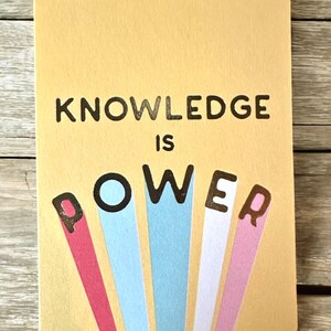 K&ouml;nnte beinhalten: Eine rechteckige Karte mit abgerundeten Ecken, die die Worte "KNOWLEDGE IS POWER" in schwarzen und farbigen Buchstaben zeigt. Das Wort "POWER" hat farbige Strahlen, die sich von jedem Buchstaben aus erstrecken, auf hellgelbem Hintergrund.