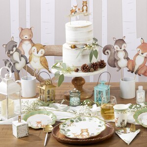 May include: A woodland-themed party table setting with a two-tiered cake, paper plates, and decorative animal cutouts. The cake is on a white stand, and the plates feature forest animal illustrations. The color scheme includes white, green, and brown.