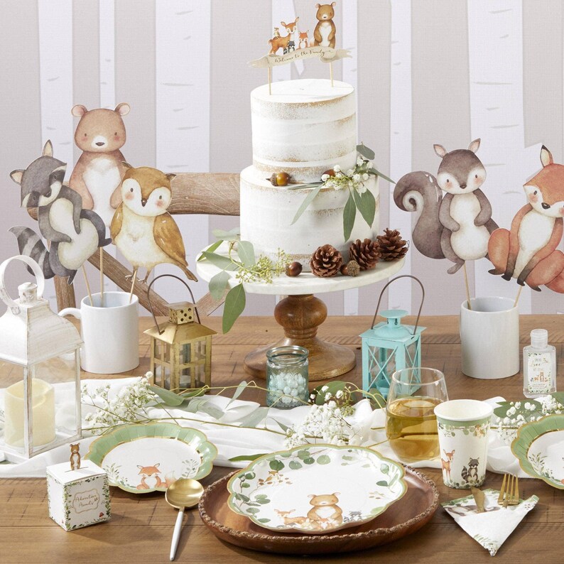 May include: A woodland-themed party table setting with a two-tiered cake, paper plates, and decorative animal cutouts. The cake is on a white stand, and the plates feature forest animal illustrations. The color scheme includes white, green, and brown.