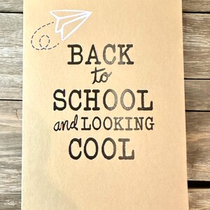 K&ouml;nnte beinhalten: Ein rechteckiges, beigefarbenes Notizbuch mit dem Text "BACK to SCHOOL and LOOKING COOL" in schwarzer Schrift. Eine wei&szlig;e Papierflugzeug-Grafik befindet sich in der oberen linken Ecke. Das Notizbuch liegt auf einer Holzoberfl&auml;che.