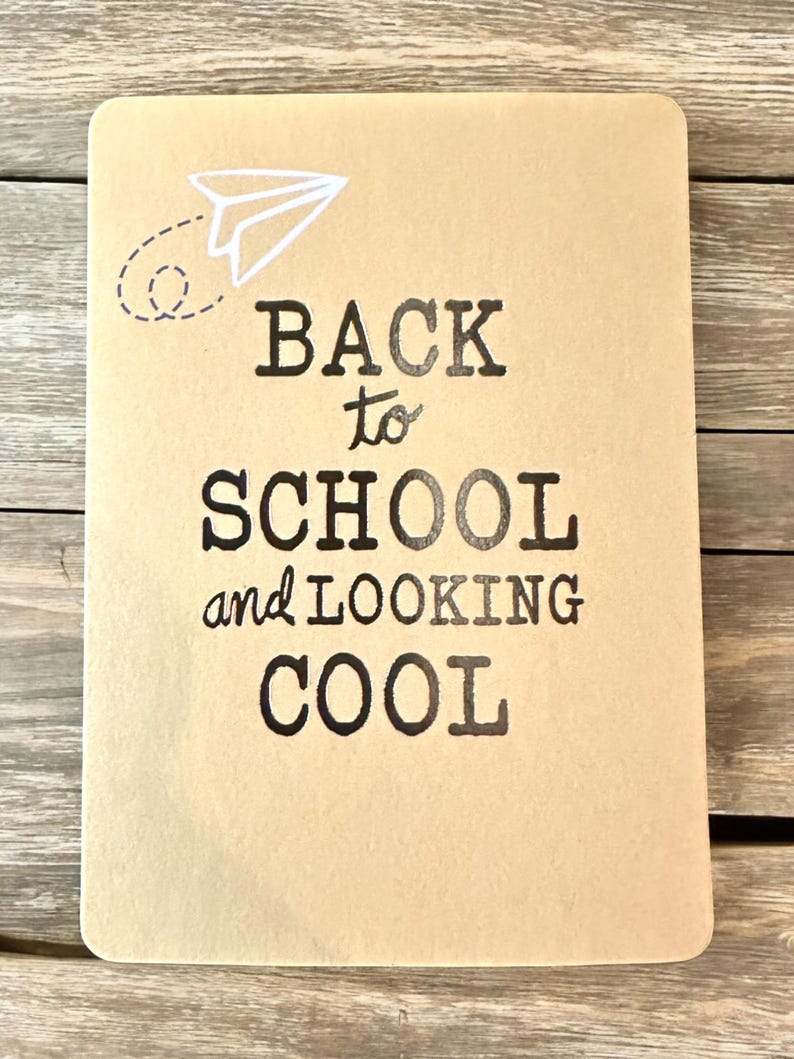 K&ouml;nnte beinhalten: Ein rechteckiges, beigefarbenes Notizbuch mit dem Text "BACK to SCHOOL and LOOKING COOL" in schwarzer Schrift. Eine wei&szlig;e Papierflugzeug-Grafik befindet sich in der oberen linken Ecke. Das Notizbuch liegt auf einer Holzoberfl&auml;che.