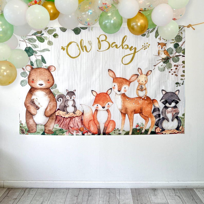 May include: A woodland-themed backdrop with the text "Oh Baby" in gold script. The backdrop features illustrations of a bear, fox, deer, raccoon, and other forest animals. Balloons in gold, white, and green are suspended above the backdrop.