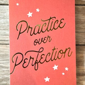 K&ouml;nnte beinhalten: Eine korallenfarbene Karte mit abgerundeten Ecken zeigt den goldfarbenen Text "Practice over Perfection". Wei&szlig;e Sterne sind &uuml;ber die Karte verstreut. Die Karte liegt auf einem Holzgrund.