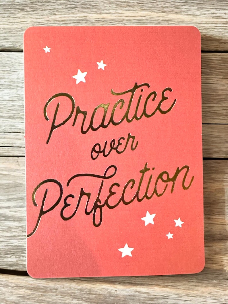 K&ouml;nnte beinhalten: Eine korallenfarbene Karte mit abgerundeten Ecken zeigt den goldfarbenen Text "Practice over Perfection". Wei&szlig;e Sterne sind &uuml;ber die Karte verstreut. Die Karte liegt auf einem Holzgrund.